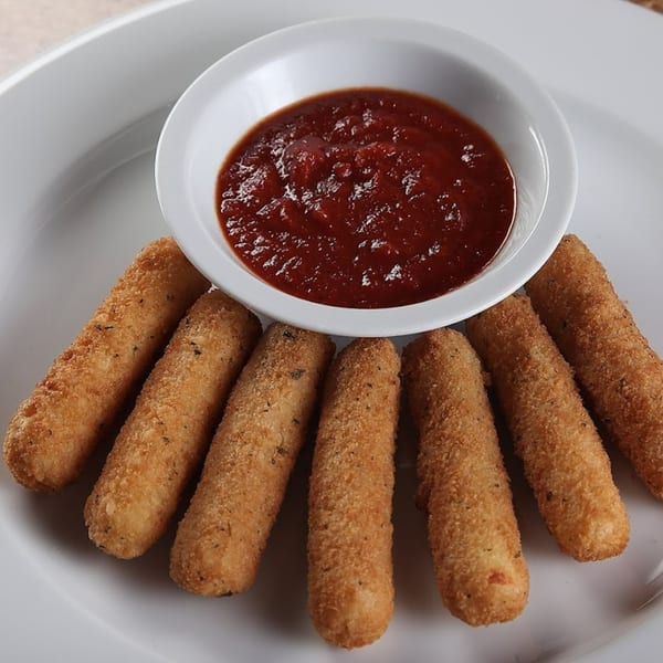 Six golden-brown mozzarella sticks with herbs, fanned around a white bowl of marinara sauce on a white plate.
