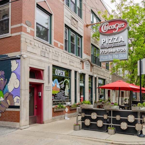 Exterior of Chicago's Pizza restaurant with red brick, large windows, and outdoor seating under red umbrellas.