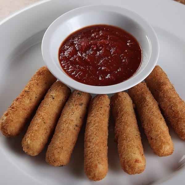 Seven golden-brown mozzarella sticks with herbs, arranged around a white bowl of marinara sauce on a white plate.