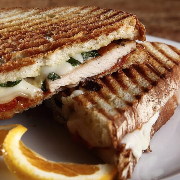 Grilled panini sandwich with chicken, melted mozzarella, basil, and tomato, served with an orange slice.