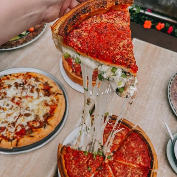Hand lifting a slice of Chicago's deep-dish pizza, with cheese stretching, surrounded by other pizzas.
